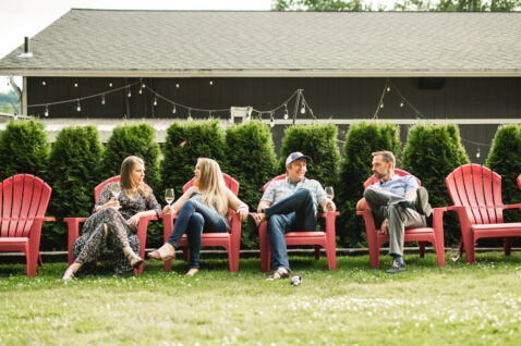Four young women and men sitting in red Adirondack chairs on the lawn socializing and drinking glasses of wine