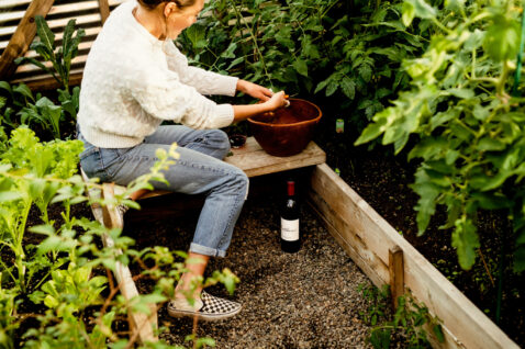 Young woman in a white seater sitting on a bench in a greenhouse picking produce and adding to a bowl