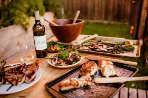 Closeup of a table full of grilled veggies, grilled chicken, salad and a bottle of Sauv Blanc