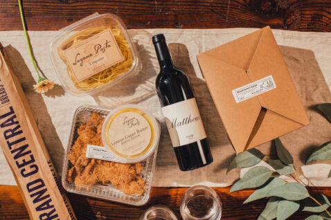 Looking down on containers of pasta, chicken, a baguette, to-go box and a bottle of Claret