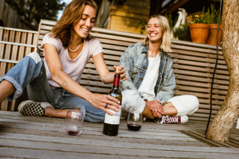 Two young woman sitting on a deck smiling and re-corking a bottle of Claret