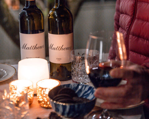 Close up of table with Matthews bottles, candles, full glass of red and a man in a red jacket