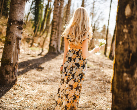 The back of a blonde female walking through the birch trees holding a glass of white wine