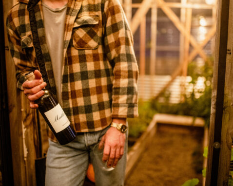 Man in flannel shirt standing at the entrance to a greenhouse holding a bottle of Claret and a stemless wineglass
