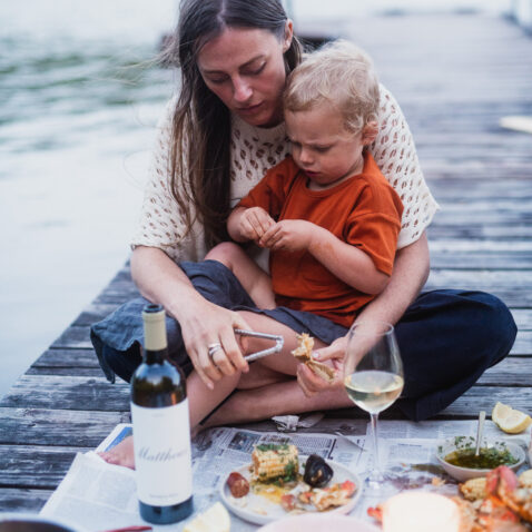 Woman with child in her lap sitting on a dock with a seafood boil spread out and Matthews Reserve Sauv Blanc