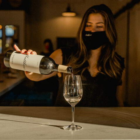 Matthews employee behind bar wearing a black mask pouring white wine in glass