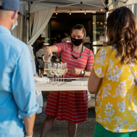 Matthews employee behind bar outside wearing a black mask, pouring white wine into multiple glasses