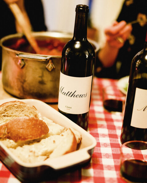 Close up of checkered red table cloth, pan of garlic bread, pot of pasta and a bottle of Claret