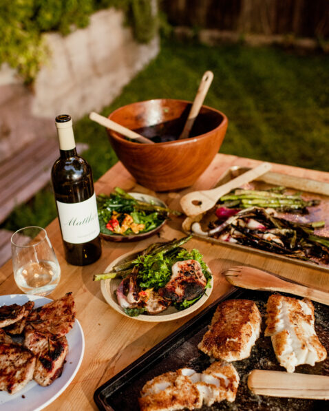 Closeup of a tablescape of grilled veggies and chicken, salad and a bottle of Sauv Blanc