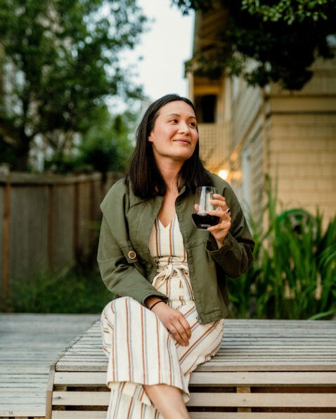 A young woman in a green jacket sitting on a bench drinking a glass of red out of a stemless wine glass