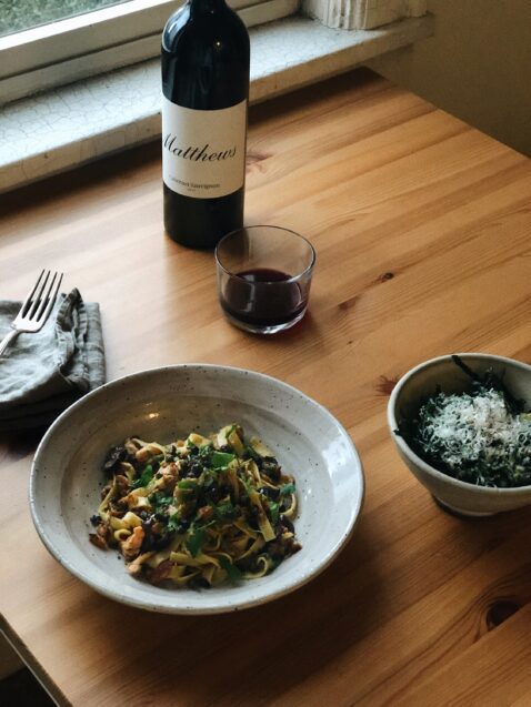A bowl of pasta, silverware, stemless glass and bottle of Cab Sauv on a table