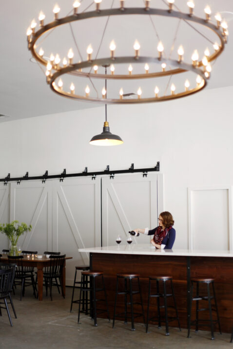A women behind the Matthews bar pouring two glasses of red, stools lines up and a two tier chandelier