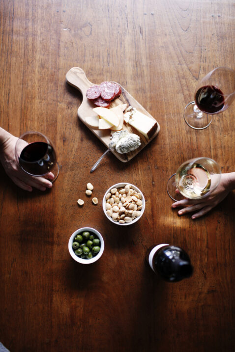Looking down on a wood table with full wine glasses, a bottle, charcuterie board, olive and almond snacks