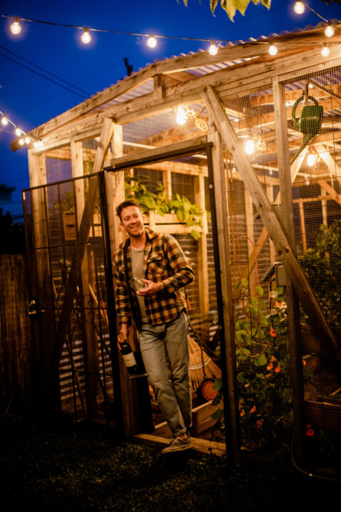 A smiling male leaning against the door of a greenhouse, under string lights and dusk sky, holding a bottle of Claret