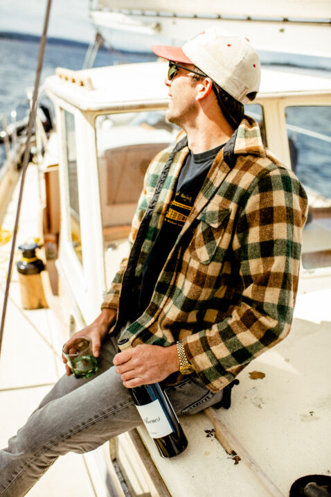 A man in a flannel and hat sitting on a sailboat, smiling and looking off into the distance holding a bottle of Sauv Blanc