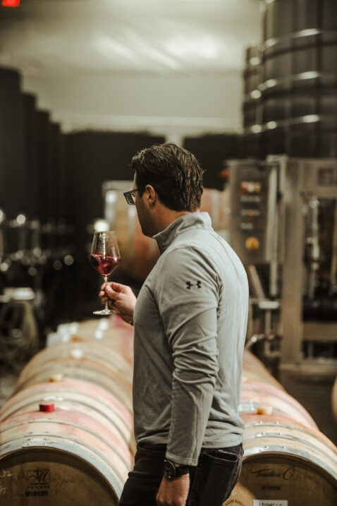 Winemaker Aryn Morell swirling a glass of red wine with a row of barrels in the background