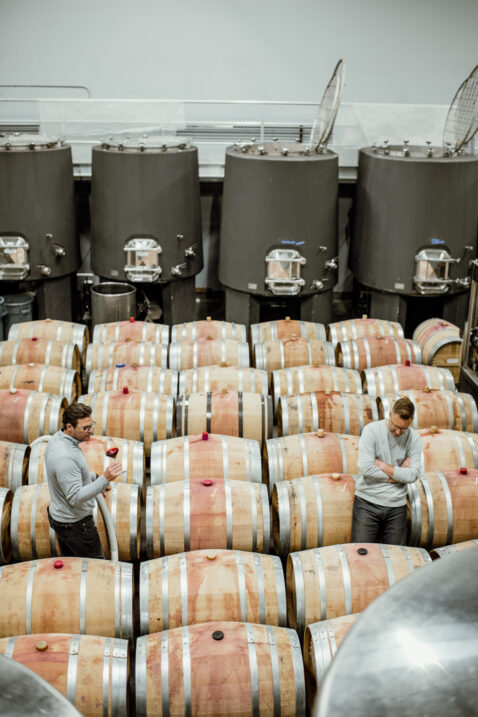Looking down on the floor of the productions facility with a row of cement tanks, rows of barrels and winemaker Aryn and owner Bryan Otis tasting