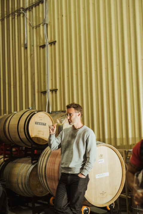 Owner Bryan Otis sniffing a glass of white wine leaning against a barrel