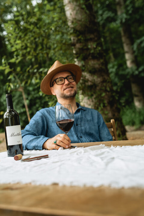 Gentleman in denim shirt and beige hat swirling a glass of red wine at a table next to a bottle of 2018 Cuvee
