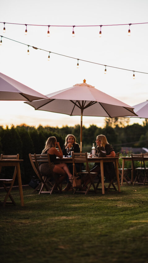 3 women sitting at a wood table under an umbrella on the upper lawn at dusk