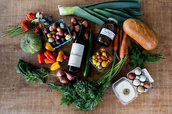 Looking down at a display of farm box items on a table, including tomatoes, a pumpkin, green produce, herbs jam, and a bottle of wine