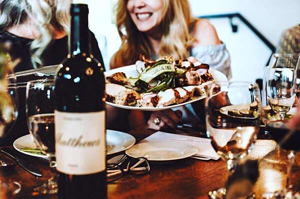 A bottle of Matthews on a dining table in the foreground, and a plate of chicken being passed to a smiling women in the background