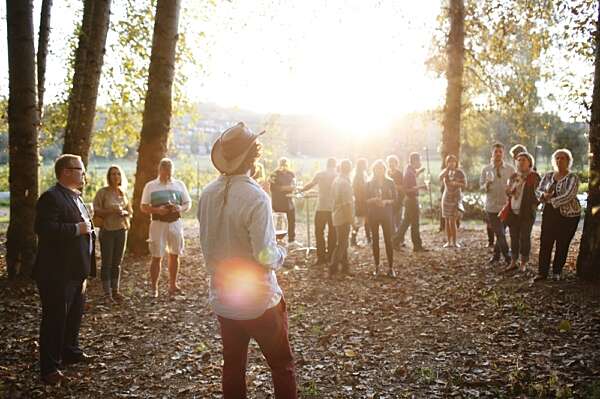 The back of a farmer in a brimmed hat speaking to a crowd of guests under the birch trees