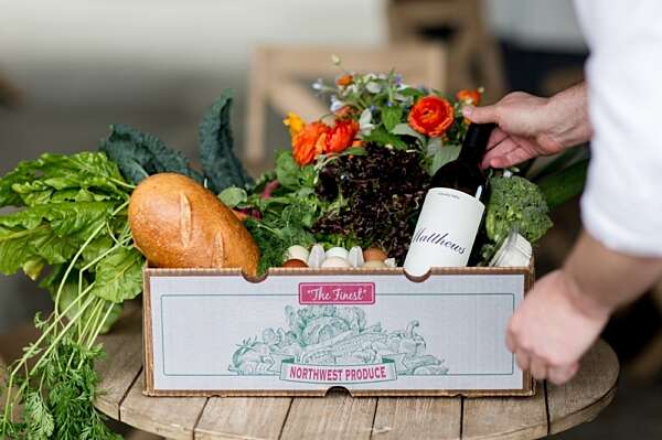 A farm box on a table stuffed with green produce, herbs, bread, eggs, flowers and a bottle of Matthews wine
