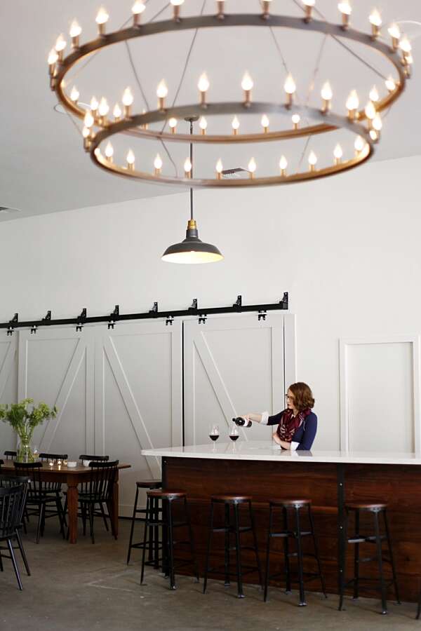 A women behind the Matthews bar pouring two glasses of red, stools lined up in front and a two tiered chandelier hanging from the ceiling
