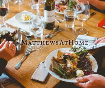 Closeup of a tablescape full of plates of food, wine glasses and a Matthews bottle, with a # matthewsathome title