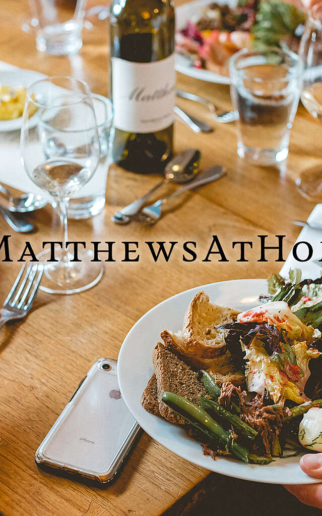 Closeup of a tablescape full of plates of food, wine glasses and a Matthews bottle, with a # matthewsathome title