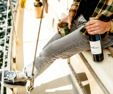 A man on the edge of a sailboat holding a bottle of Sauv Blanc and an emerald green glass