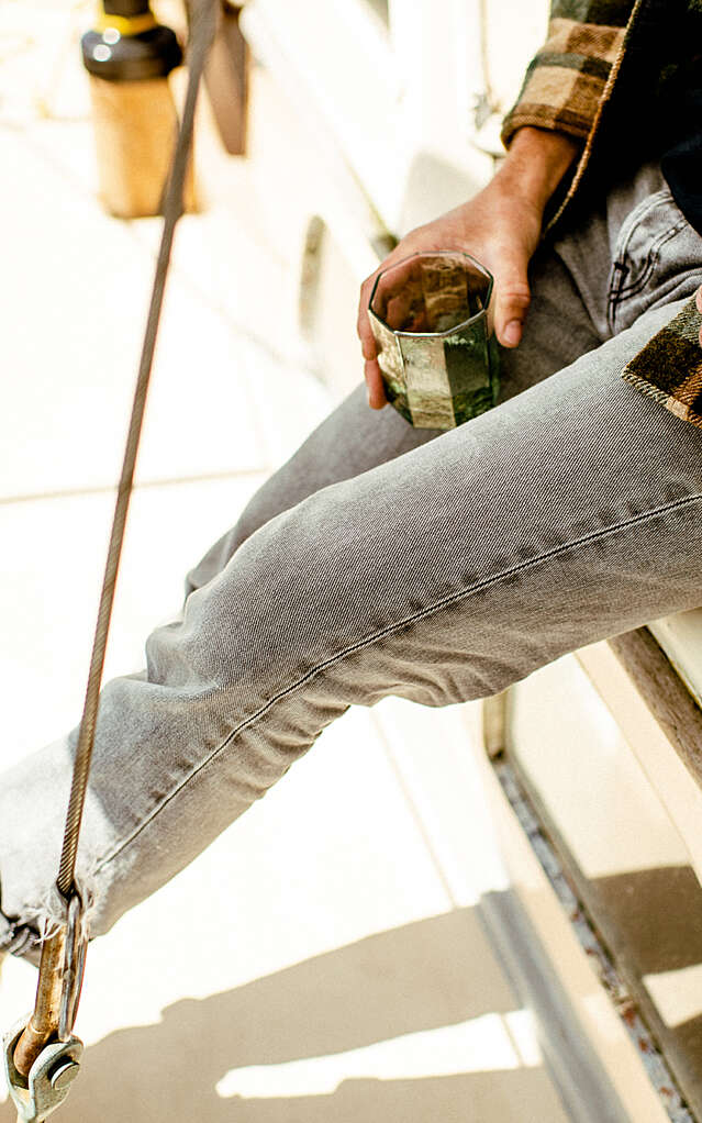 A man on the edge of a sailboat holding a bottle of Sauv Blanc and an emerald green glass