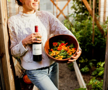 Young woman in white sweater standing in the entrance to a greenhouse, holding a bottle of Claret and a bowl of florals