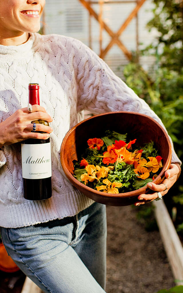 Young woman in white sweater standing in the entrance to a greenhouse, holding a bottle of Claret and a bowl of florals